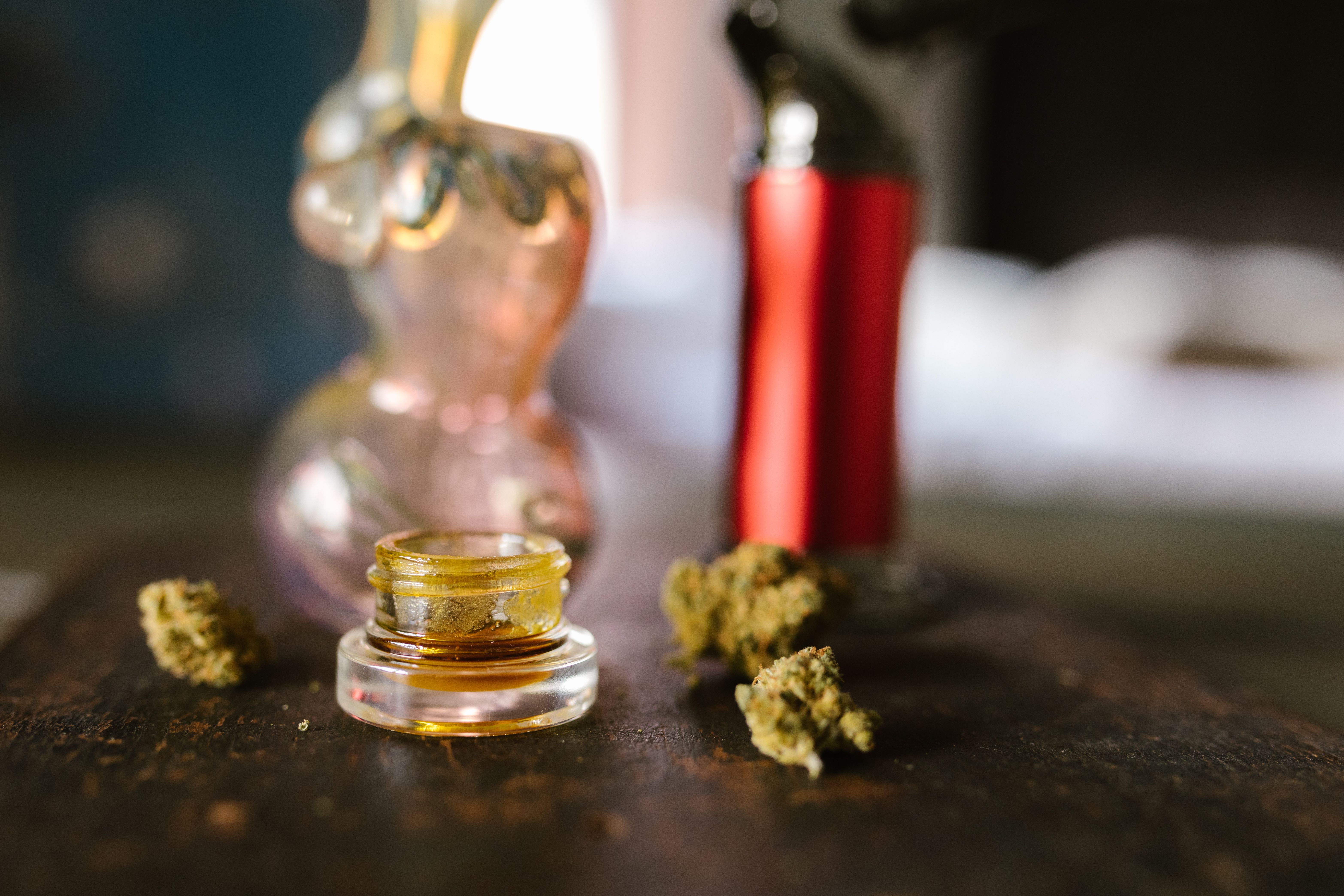 Glass jar of cannabis concentrate with dried buds and smoking accessories on a table
