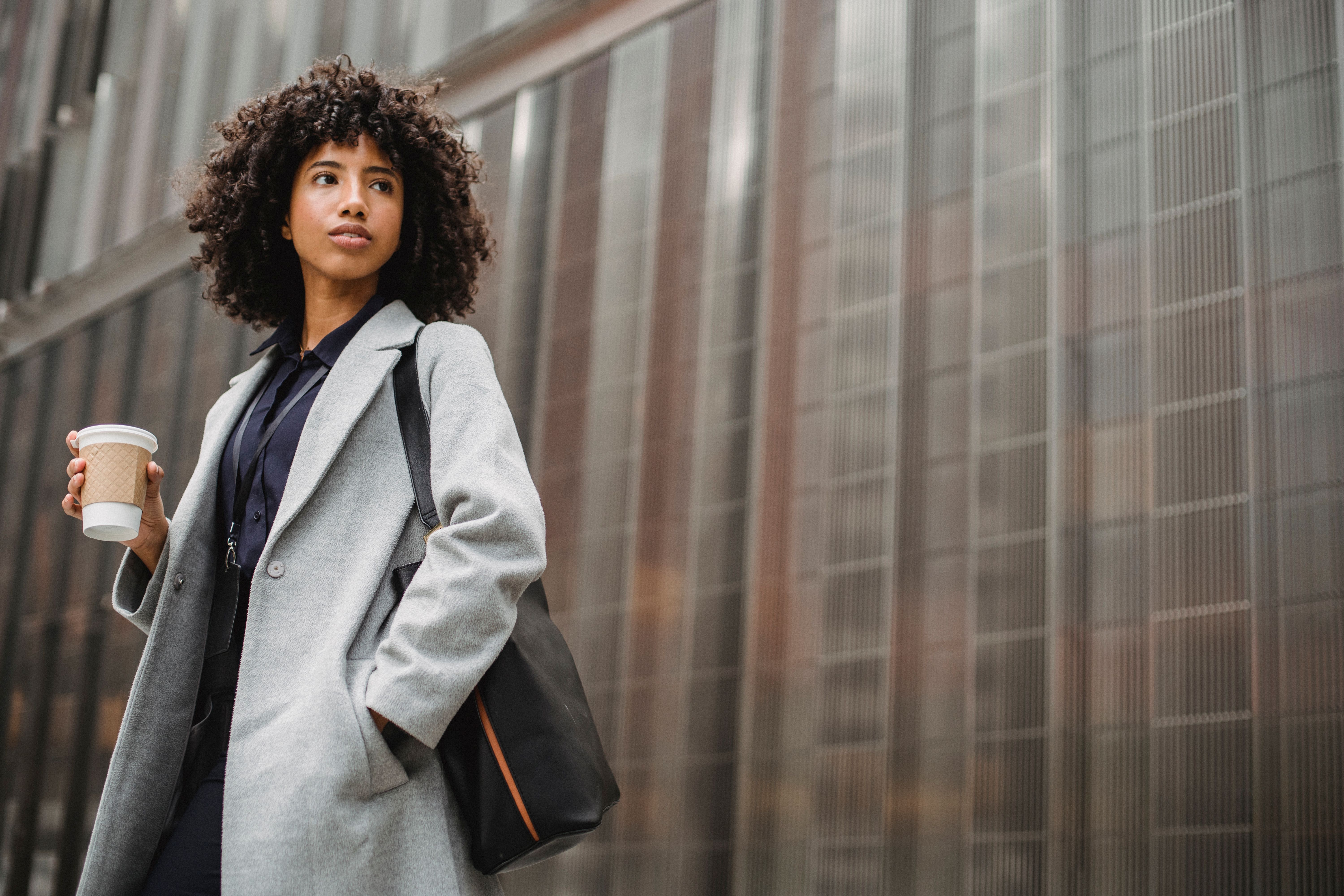 Confident woman in a gray coat standing poised against a modern city building